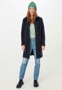 Abrigo de mezcla de lana azul marino con botones, llevado sobre una camisa a rayas verde y blanca, combinado con jeans azul claro y botas negras de tobillo.