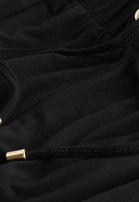 Tela negra con pliegues horizontales, con acentos de hardware en tono dorado y detalles trenzados a lo largo de los bordes. Material suave y texturizado.