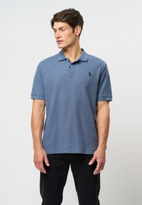 Svetlomodrá bavlnená polokošeľa s klasickým golierom, krátkymi rukávmi a malým čiernym logom na hrudi. Nosí sa s čiernymi nohavicami.