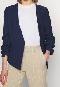 Navy blue blazer with a relaxed fit and rolled sleeves, worn over a plain white t-shirt and paired with beige striped trousers.