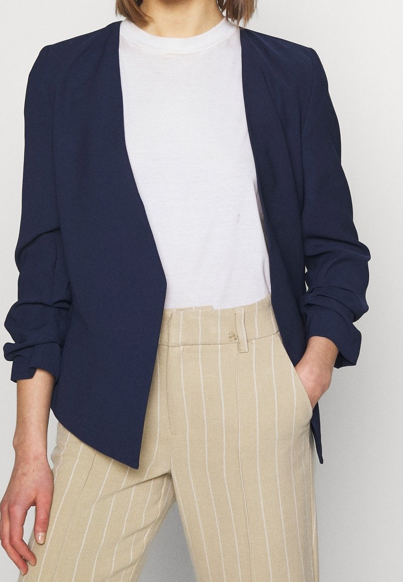 Navy blue blazer with a relaxed fit and rolled sleeves, worn over a plain white t-shirt and paired with beige striped trousers.