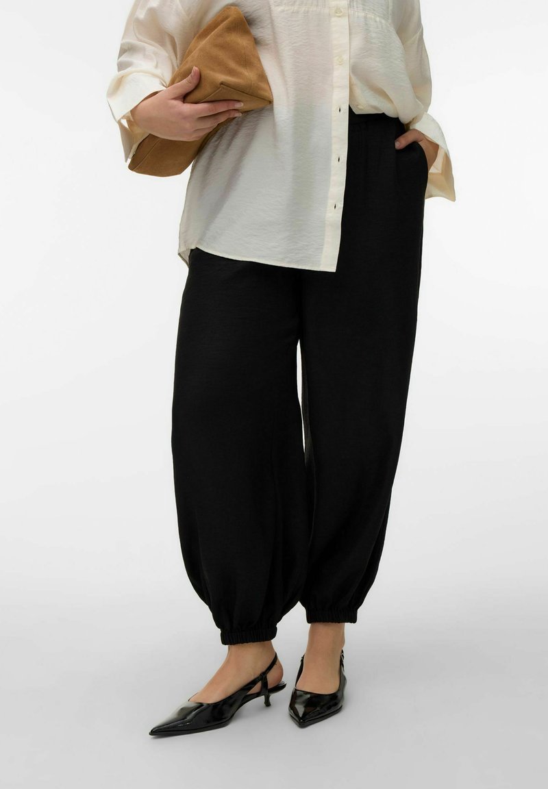 Femme portant un pantalon noir ample avec revers, une chemise crème à boutons, des escarpins noirs à bride arrière, et tenant une pochette beige sous un bras.