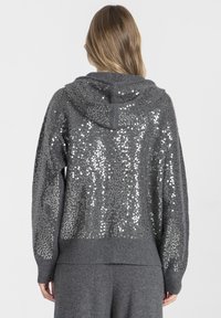 Szara bluza z kapturem z tyłem ozdobionym cekinami, teksturowanym dzianinowym materiałem oraz ściągaczami na dole i przy mankietach. Luźny, oversize'owy krój.