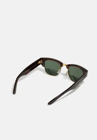 Kilpkonnakilp sunglasses roheliste läätsedega, kuldsete aktsentidega, nurgelise raami ja lamedate tempidega. Omab nina padjakesi ja allkirja detaile.