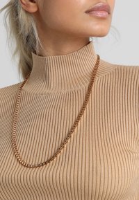 Collier chaîne en métal doré présenté sur un pull à col montant beige côtelé, mettant en valeur sa texture lisse et ses maillons géométriques.