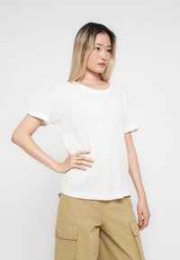 Young woman with blonde ombre hair wearing a white t-shirt and tan cargo pants, standing with one hand on her hip against a white background.