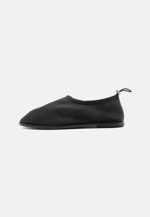 Zapato de tela negro tipo slip-on con suela plana y lazo elástico ajustable en el talón, mostrado desde el lado exterior sobre fondo blanco.