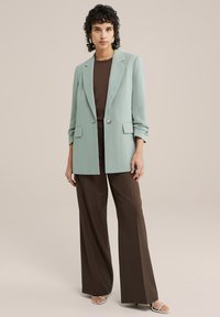 Světle zelený blazer se strukturovaným designem, jednoduchým zapínáním na knoflík a předními kapsami, spárovaný s hnědými širokými kalhotami a sandály.