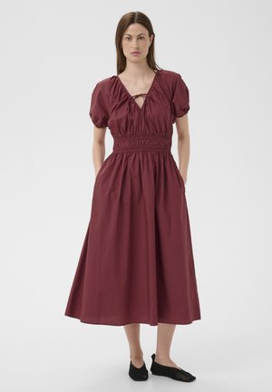 Femme debout portant une robe midi bordeaux avec des manches bouffantes, une taille smockée, un col en V avec un détail de lien, et des chaussures noires sans lacets.