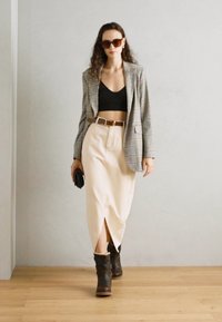Blazer à carreaux, top court noir, jupe beige taille haute avec fente, ceinture marron, bottines, lunettes de soleil, tenant une pochette noire.