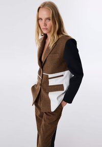 Bruine geruite blazer met tonale accenten, met een witte gestreepte sectie en zwarte mouwen. Gepersonaliseerde pasvorm met klepzakken.