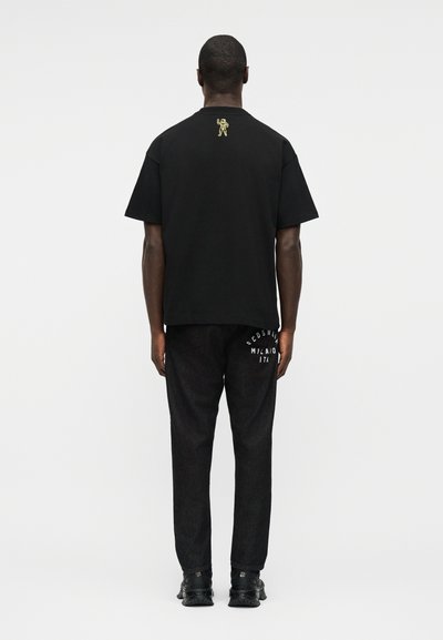 Άνδρας που φορά μαύρο t-shirt με μικρό χρυσό αστροναύτη στο πάνω μέρος της πλάτης και μαύρο παντελόνι με λευκό κείμενο στην περιοχή της δεξιάς τσέπης.