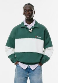 Felpa a blocchi di colore verde e bianco con colletto, dotata del testo "STWD", indossata sopra una camicia a righe chiara. Texture morbida.