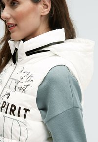 Weiße, gepolsterte Weste mit Kapuze, die schwarze Akzente und Textdetails aufweist, kombiniert mit einem hellblauen Pullover aus strukturiertem Stoff.