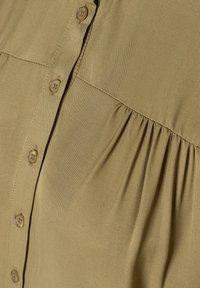 Haut khaki à boutons avec un tissu texturé, des détails de design plissés et six boutons ronds marron clair sur le devant.