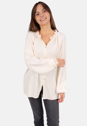 Femme souriante, portant un chemisier blanc ample à manches longues avec bordure en dentelle et un pantalon gris foncé, debout devant un fond uni.