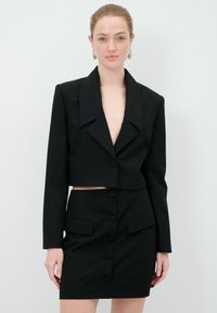 Blazer nero cropped con ampi revers e tessuto testurizzato, abbinato a una gonna alta coordinata con bottoni frontali e tasche laterali.