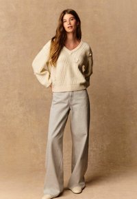 Krämfärgad stickad v-ringad tröja med texturerade mönster, kombinerad med ljusgrå vida jeans och beigea slip-on skor. Slät, avslappnad look.