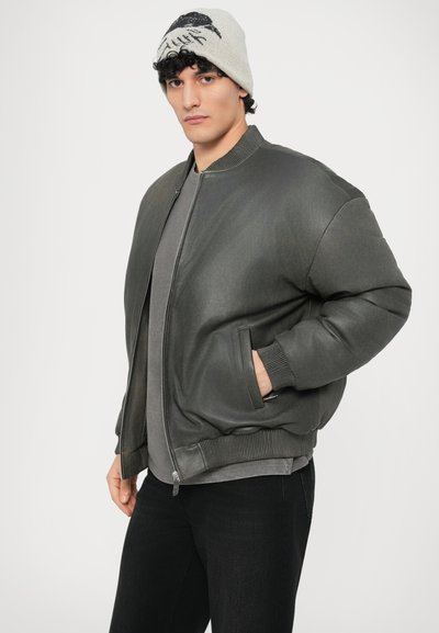 Tamsiai pilkos spalvos bomberio stiliaus striukė iš lygaus audinio, su apsiūtais rankogaliais, dviem šoniniais kišenėmis ir užtrauktuku. Dėvima ant pilkos spalvos marškinėlių.