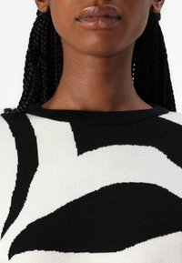 Mustavalkoinen neule, jossa on rohkea abstrakti kuvio, ribatty kaulus ja pehmeä neulostekstuuri, näkyvillä ompeluyksityiskohdilla.