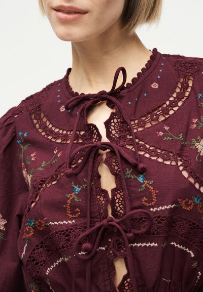 Κοντινό πλάνο ενός ατόμου που φοράει μπλούζα μπορντό με κεντήματα λουλουδιών, δαντελωτές εξοχές και τρία δεμένα φιόγκια στο λαιμό.