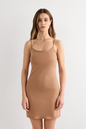 Donna con capelli lunghi castano chiaro che indossa un vestito beige aderente, senza maniche e lungo fino al ginocchio, in piedi davanti a uno sfondo bianco semplice.