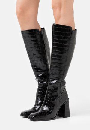 Glänzende schwarze kniehohe Stiefel mit Krokodillederstruktur, Blockabsätzen und seitlichen Reißverschlüssen, getragen an nackten Beinen.