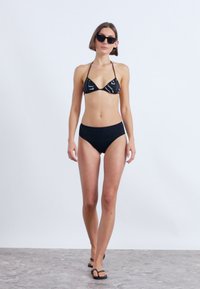 Μαύρο μπικίνι σετ με τριγωνική κορυφή με λευκές σχέσεις και ψηλόμεσα σορτς. Το μοντέλο φορά μαύρα γυαλιά ηλίου και σαγιονάρες.