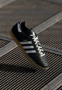 Juodos odinės sportinės bateliai su baltais dryžiais ir auksiniu "SAMBA" tekstu. Yra guminis padas ir paminkštinta apykaklė patogumui. Pasirinkti klasikiniai dizaino elementai.