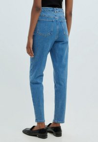 Persona que lleva un pantalón vaquero azul de tiro alto y mocasines negros, de pie con una mano relajada a su lado frente a un fondo liso.