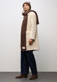 Krämsk faux fur-kappa med brun etikett, brun stickad halsduk, blå vida jeans och bruna ankelboots. Mot en ljus bakgrund.