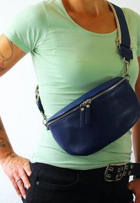 Donkerblauwe leren heuptas met een gestructureerd oppervlak, voorzien van een bovenrijpsluiting en een verstelbare band, gedragen om de taille in een casual setting.