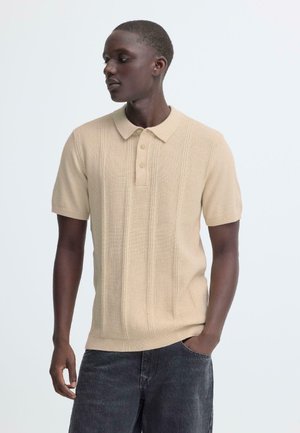 Polo beige à manches courtes avec un motif en maille texturée, patte à deux boutons et col côtelé, porté avec un jean foncé.