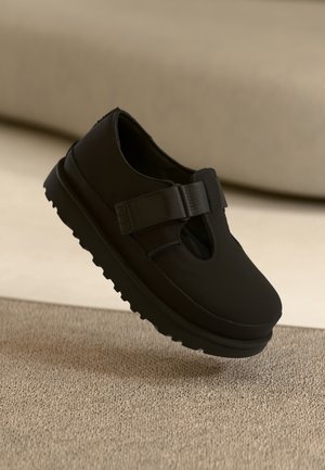 Musta liukuva kenkä, jossa on sileä pinta, pyöreä kärki, joustava nauha ja paksu, uurrettu pohja paremman pidon takaamiseksi.
