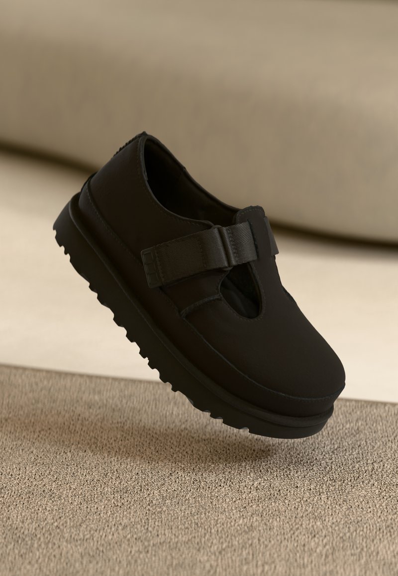 Sapato preto tipo slip-on com textura suave, apresentando um bico redondo, uma tira elástica e uma sola grossa e estriada para maior aderência.