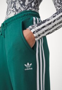 Osoba nosząca zielone spodnie dresowe Adidas z białymi paskami i logo, z ręką w kieszeni, w połączeniu z czarno-białą wzorzystą bluzką z długim rękawem.