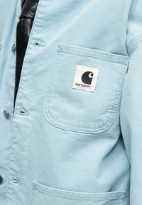 Ελαφρώς γαλάζιο μπουφάν με εμφανείς μπροστινές τσέπες, μαύρα κουμπιά και μαύρο-λευκή ετικέτα λογότυπου Carhartt στην επάνω τσέπη.