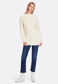 Chunky knit cream sweater with textured upper and ribbed lower section, paired with blue jeans and white ankle boots.