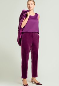 Haut sans manches en satin violet, assorti à un pantalon en velours violet. Le mannequin tient un blazer assorti, porte des escarpins vernis bordeaux.