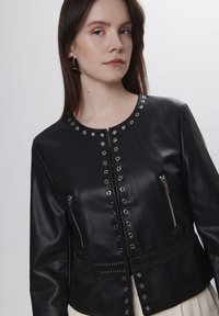 Giacca in pelle nera con borchie in metallo argentato, tasche laterali con zip e un dettaglio di cintura intrecciata sulla vita. Finitura liscia e design aderente.