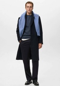 Cappotto di lana navy con una vestibilità su misura, accompagnato da una sciarpa di maglia azzurro chiaro. I pantaloni e le scarpe neri completano l'outfit. Texture complessivamente liscia.