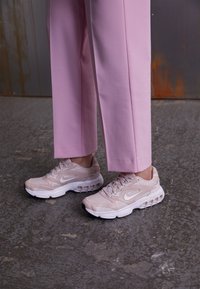 Sneakers Nike rosa chiaro con tomaia in mesh, dettagli bianchi e suola imbottita. Abbinati a pantaloni rosa su misura su una superficie grigia texturizzata.