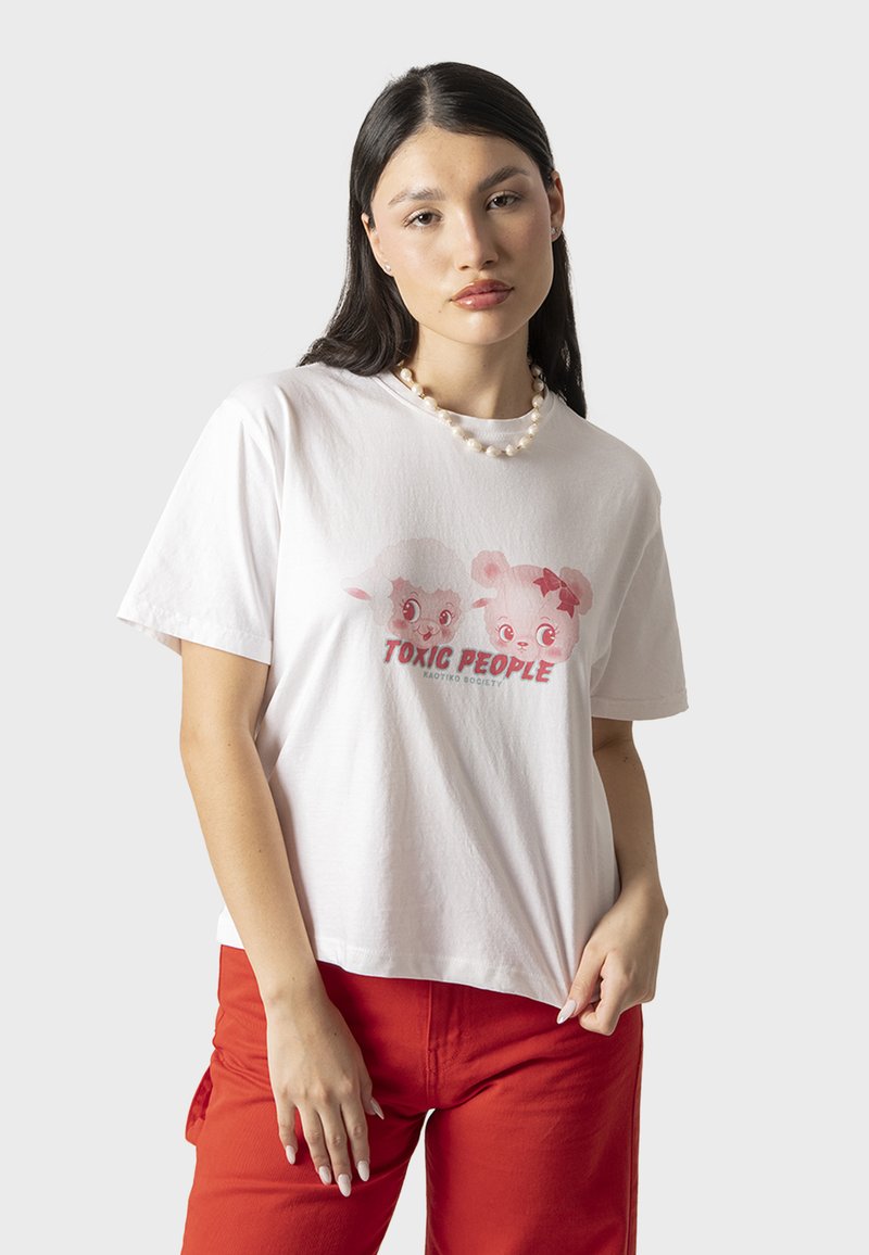 Witte katoenen t-shirt met een grafiek van twee cartoonfiguren en de tekst "TOXIC PEOPLE" eronder. Model draagt rode broek en parelketting.