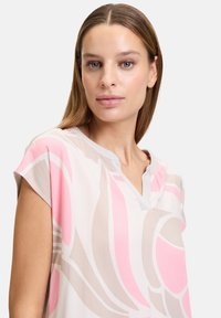 Bluzka z krótkim rękawem z lekkiego materiału w różowo-beżowy wzór kwiatowy, z dekoltem w kształcie litery V i szarymi akcentami kołnierza.