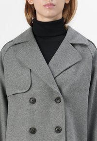 Cappotto grigio doppiopetto in tessuto di lana testurizzata, con ampi risvolti e bottoni neri, indossato sopra un dolcevita nero.