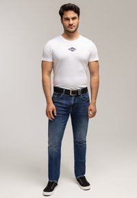 Młody mężczyzna stoi, ubrany w biały t-shirt z grafiką, niebieskie dżinsy, czarny pasek i czarne sportowe buty na jednolitym jasnym tle.