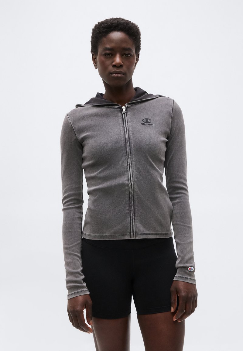 Femme portant un sweat à capuche gris zippé avec logo et un short noir, debout devant un fond clair uni.