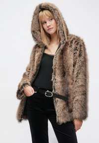 Pruunist kunstkarvast kapuutsiga jakk luku ja rikkaliku tekstuuriga, kontrastse tumeda vööga, kombineeritud musta pluusi ja pükstega.