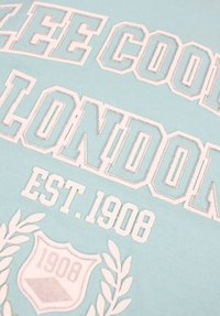 Tissu en coton bleu clair avec un texte blanc audacieux indiquant "LEE COOPER LONDON" accompagné de la date de création "EST. 1908" et d'un motif de laurier.