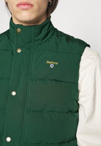 Muž v tmavě zelené prošívané vestě Barbour přes světle béžovou košili s dlouhým rukávem, viditelné klopy, knoflíky a logo na hrudi.